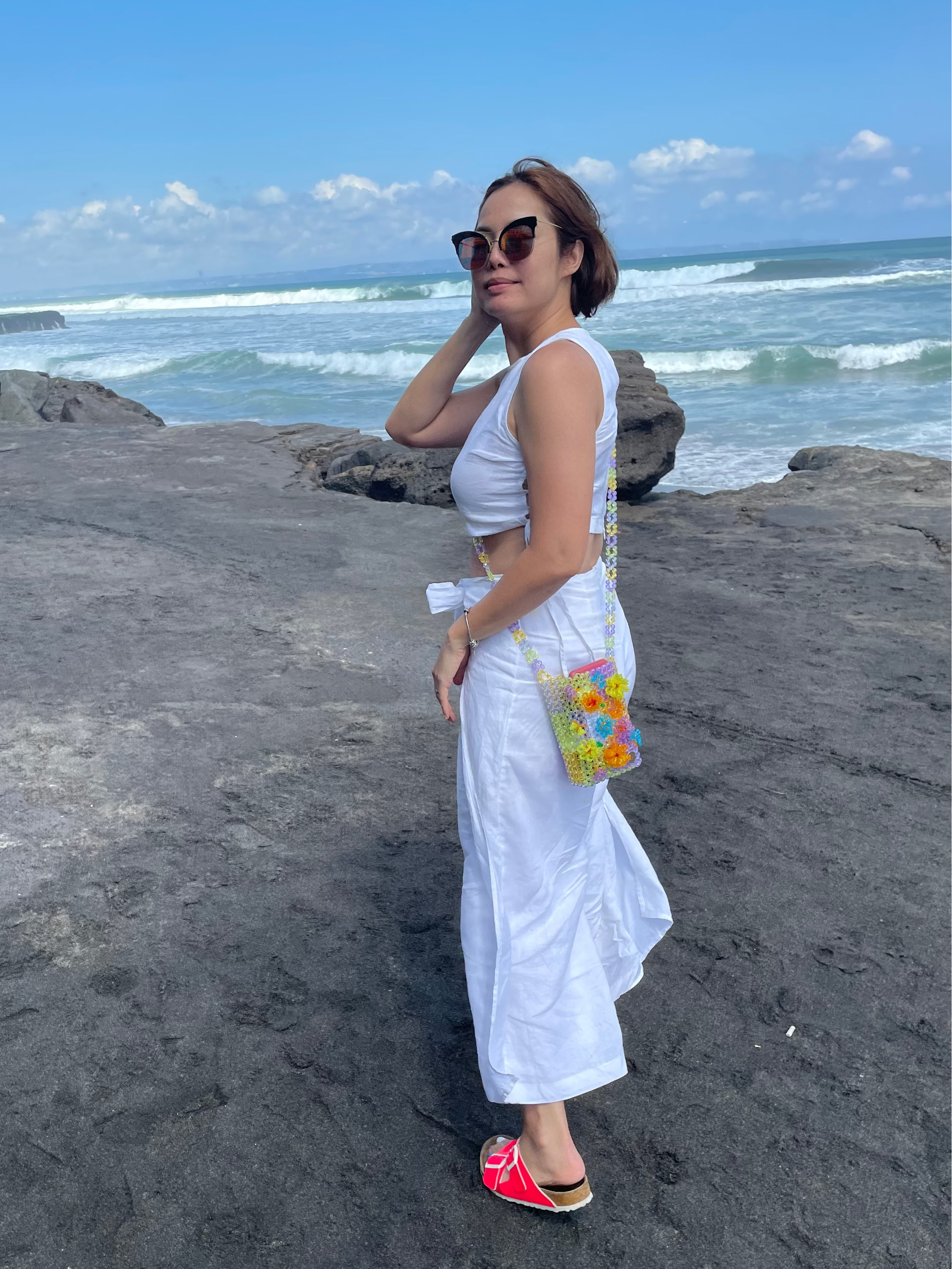 Tern posing on a black volcanic rock beach wearing a white two-piece crop top and maxi skirt set, a small colorful floral crossbody bag, and pink sandals, with the ocean and blue sky in the background.