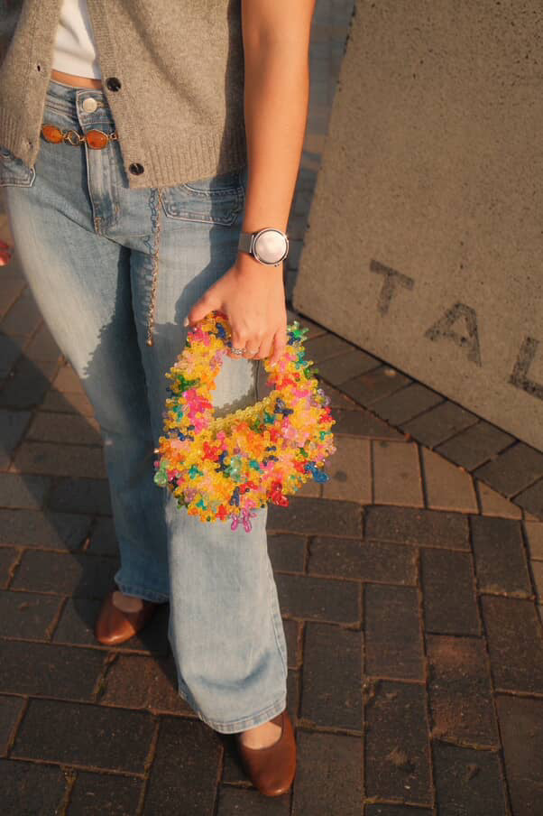 A person outdoors holding a small, colorful, flower-like handbag with a textured, bubbly surface. The person is wearing light-wash blue jeans, a beige cardigan, and brown leather shoes, standing on a dark brick pavement next to a concrete wall.