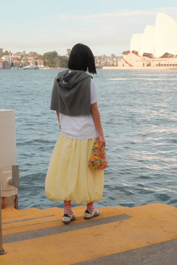 A person standing on a pier by the water, seen from behind, wearing a white T-shirt, a long pleated light yellow skirt, and a dark gray hoodie draped over their shoulders. The Sydney Opera House is visible in the background across the water.