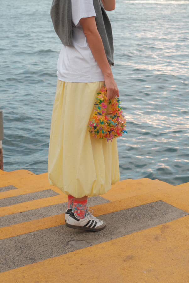 A person standing on a yellow pier with water in the background, wearing a white t-shirt, a long pale yellow skirt, a gray cardigan, colorful socks, and white sneakers, while holding a small multicolored floral bag.
