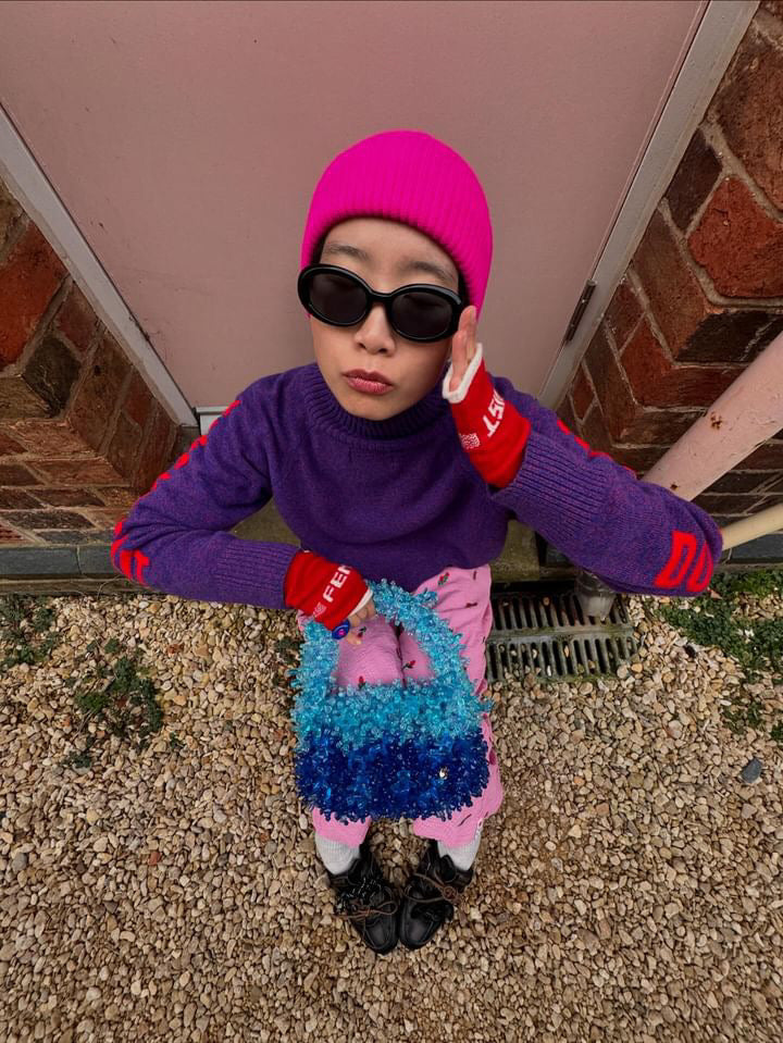 She wearing a colorful outfit with a bright pink hat and sunglasses, standing on a gravel surface.