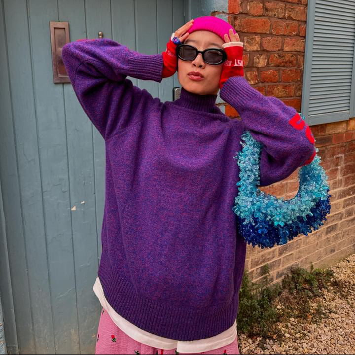 Person wearing a purple sweater and pink headscarf holding a colorful wreath against a brick wall.