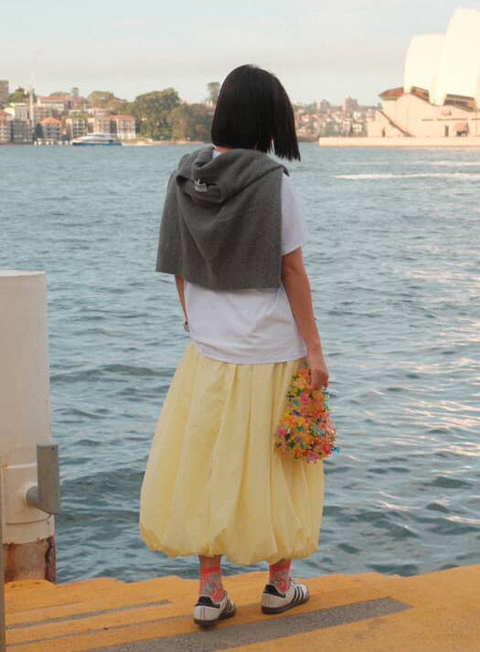 A person standing on a pier by the water, seen from behind, wearing a white T-shirt, a long pleated light yellow skirt, and a dark gray hoodie draped over their shoulders. The Sydney Opera House is visible in the background across the water.